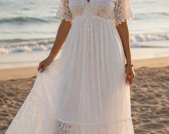 May include: A white maxi dress featuring a V-neckline, short lace sleeves, and a lace bodice. The dress has a drawstring waist and a long, flowing skirt with a tiered lace panel at the bottom hem, crafted from a combination of lace and fabric.