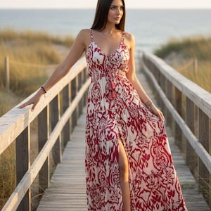 Puede incluir: Un vestido largo hasta los pies con un estampado de motivos en rojo oscuro y blanco. La prenda tiene tirantes finos, escote en V, cintura ceñida con un lazo decorativo de borlas y un diseño de falda escalonada con una abertura frontal.