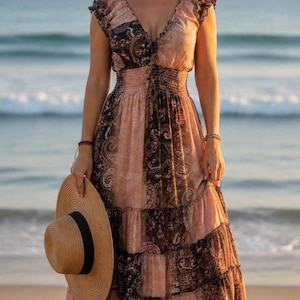 May include: A floor-length maxi dress featuring a v-neckline, ruffled cap sleeves, and a smocked waist. The garment is crafted from a lightweight fabric with a dark brown and peach paisley print, tiered skirt panels, and a wide-brimmed straw hat.
