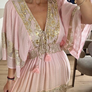 Pu&ograve; includere: Un abito rosa chiaro con ampie maniche a volant, scollo a V profondo e vita elasticizzata con coulisse e nappe. Il capo &egrave; decorato con intricati ricami metallici dorati e motivi floreali lungo il corpetto e la gonna.