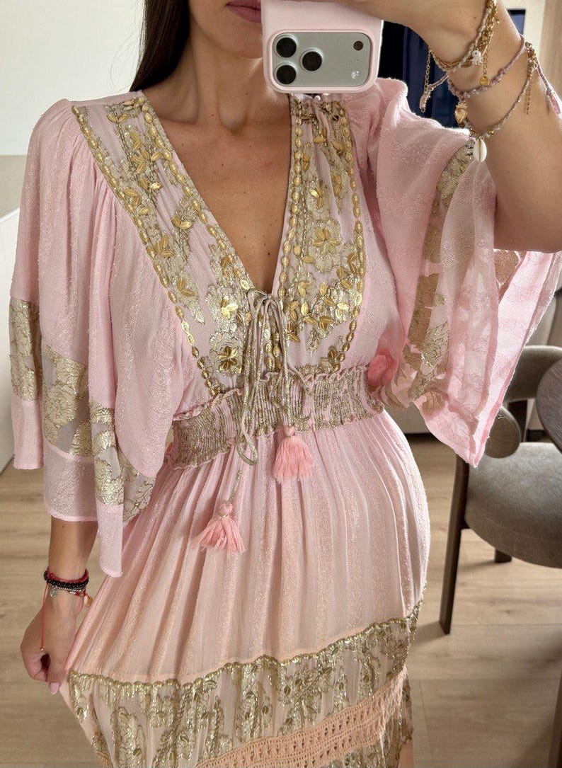 Pu&ograve; includere: Un abito rosa chiaro con ampie maniche a volant, scollo a V profondo e vita elasticizzata con coulisse e nappe. Il capo &egrave; decorato con intricati ricami metallici dorati e motivi floreali lungo il corpetto e la gonna.