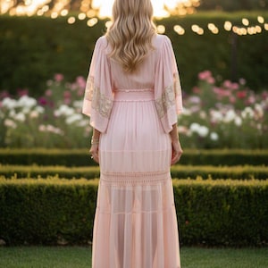Pu&ograve; includere: Un abito lungo color rosa pallido con ampie maniche a kimono impreziosite da dettagli in pizzo dorato, una vita arricciata e una gonna a balze con inserti in pizzo orizzontale. Il capo presenta una silhouette fluida e semitrasparente.
