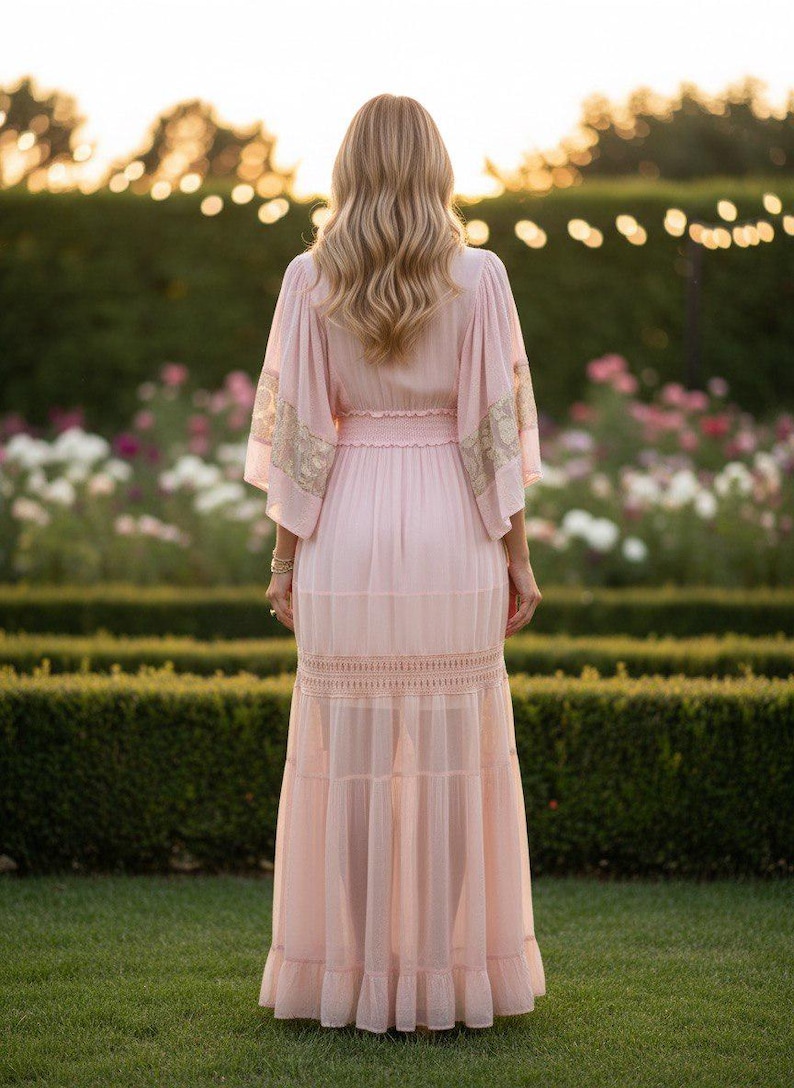 Pu&ograve; includere: Un abito lungo color rosa pallido con ampie maniche a kimono impreziosite da dettagli in pizzo dorato, una vita arricciata e una gonna a balze con inserti in pizzo orizzontale. Il capo presenta una silhouette fluida e semitrasparente.