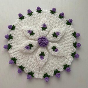 May include: A handmade, white crocheted doily with a floral design. The doily features a central flower and petal-shaped accents in purple and green. The edges are adorned with small purple buds and green leaves.