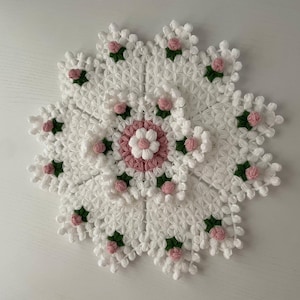 May include: A white, handmade crochet doily with a floral design. The doily features a central flower with pink and white petals, surrounded by white petals. Small pink flowers with green leaves are evenly spaced around the edges.