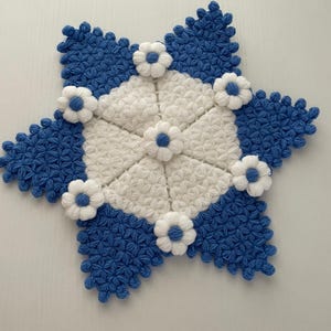 May include: A handmade, star-shaped crochet item in blue and white. The center is white, surrounded by blue points. Small white flowers with blue centers are evenly spaced around the edges. The item is likely a decorative piece or coaster.