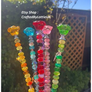 Può includere: Una collezione di paletti da giardino colorati con perline e fiori di vetro. I paletti presentano una varietà di colori, tra cui giallo, blu, rosso, rosa e verde. Il testo "Etsy Shop: CraftedByLottieUK" è visibile nell'immagine.