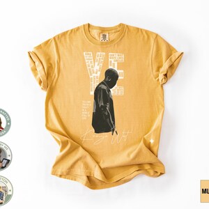 May include: Mustard-colored t-shirt featuring a black and white graphic of a person and the text "YE" and "Kanye West". The shirt also has additional text and is made of 100% cotton.
