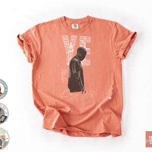 May include: Terracotta-colored t-shirt featuring a black and white image of a person and the name "Kanye West" in a handwritten font. The shirt also has the words "VE" and other text in a white font. The shirt is made of 100% cotton.