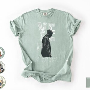 May include: A light green t-shirt featuring a black and white graphic of a person and the word "YE" in white. The shirt also has the words "Kanye West" in white script. The shirt is made of 100% cotton.