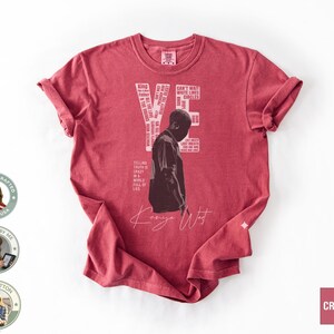 May include: A red t-shirt featuring a graphic of Kanye West with the word "YE" and other text. The shirt includes the text "AMERICAN CRAFTED MADE IN USA DESIGNED BY ME 100% COTTON" and the word "CRIMSON".