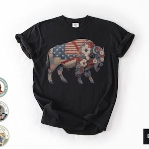 May include: Black t-shirt featuring a detailed graphic of a bison. The bison is adorned with a patchwork design incorporating the American flag and floral elements in red, white, and blue. The shirt is made of 100% cotton.