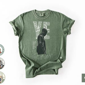 May include: Moss green t-shirt featuring a graphic of a person and the name "YE" in white block letters. The shirt also includes the text "Can't Wait" and "Circles". The words "Kanye West" are written in a cursive font. The shirt is made of 100% cotton.