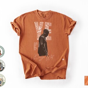 May include: Burnt orange t-shirt featuring a graphic of a person and the text "YE" with additional text. The shirt also has the signature "Kanye West" in white script. Made of 100% cotton.