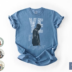 May include: Blue jean-colored t-shirt featuring a black and white image of a person. The word "YE" is printed in white, along with other text. The shirt also has the words "Kanye West" in a cursive font.