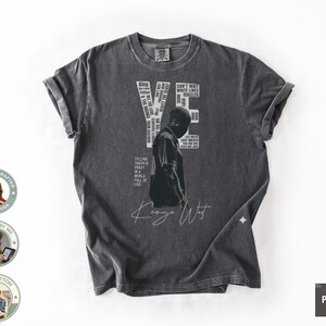 May include: Dark gray t-shirt featuring a graphic of a person and the text "YE" and "Kanye West". The shirt includes additional text elements. The shirt is made of 100% cotton.