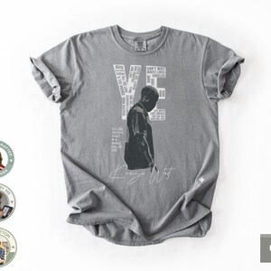 May include: A gray t-shirt featuring a graphic of a person and the word "YE" in white. The shirt includes text and the signature "Kanye West". The shirt is made of 100% cotton.