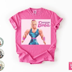 Könnte beinhalten: Ein pinkfarbenes T-Shirt mit einem Aufdruck einer muskulösen Barbie-Puppe und dem Text "Bodybuilding Barbie". Die Puppe trägt ein blaues Outfit mit pinkfarbenem Gürtel und Armbändern. Das T-Shirt hat hochgekrempelte Ärmel.
