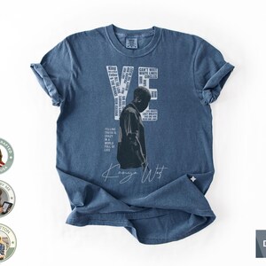 May include: Denim blue t-shirt featuring a black and white image of a person and the text "YE" with various words and phrases. The shirt also includes the text "Kanye West" in a handwritten font. Made of 100% cotton.