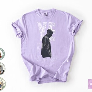 May include: A light purple t-shirt featuring a black and white image of a person and the text "YE" in white. Additional white text is arranged vertically on the right side of the image. The shirt has short sleeves and a crew neck.