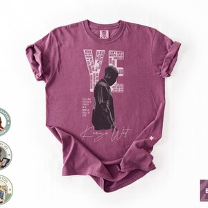 May include: Berry-colored t-shirt featuring a graphic of a person and the word "YE" in white. The shirt includes text elements and the signature "Kanye West". The shirt is made of 100% cotton.