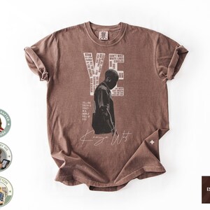 May include: Espresso-colored t-shirt featuring a black and white graphic of a person and the name "Kanye West" in a cursive font. The shirt includes text elements such as "YE" and "CAN'T WAIT WHITE LINES CIRCLES". The shirt is made of 100% cotton.