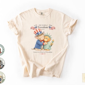 Può includere: T-shirt color avorio con grafica patriottica. Il design include un gatto dei cartoni animati con un cappello da Zio Sam che abbraccia una figura della Statua della Libertà. Il testo recita "1776 250 YEARS of 2026 Freedom" e "Love is a Kind of Freedom."