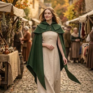 Puede incluir: Un vestido largo sin mangas de color beige claro combinado con una capa verde oscuro con capucha, que presenta un diseño de hombros superpuestos y dos botones frontales. La capa cae sobre los hombros y se extiende por los lados.