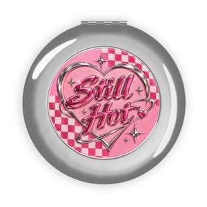 May include: A round silver compact mirror featuring a pink and white checkered background. The center displays a metallic chrome heart outline surrounding the pink chrome text Still Hot, accented by small four-pointed star and heart icons.