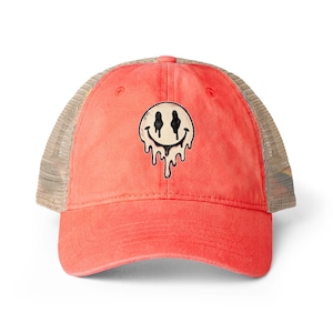 May include: A coral-colored trucker hat featuring a beige mesh back and a front panel with a graphic of a cream-colored smiley face with black eyes and a mouth that appears to be melting downward in a dripping effect against the fabric.