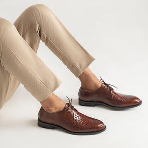 Puede incluir: Un par de zapatos oxford de cuero marrón con un patrón texturizado similar a la piel de serpiente en la puntera y los paneles laterales, con cordones finos marrones, tacón bajo y acabado de cuero liso en el talón, usados con pantalones beige.