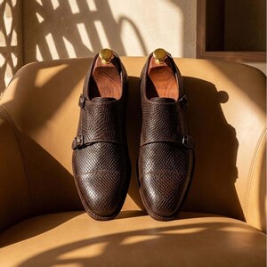 May include: A pair of dark brown double monk strap shoes featuring a textured, grid-like pattern on the leather upper. Each shoe includes two metallic buckles on the side and a wooden shoe tree inserted inside the opening of the footwear.