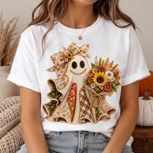 May include: A white short-sleeved t-shirt featuring a central graphic of a ghost character made of various autumn-colored floral and plaid fabric patterns. The ghost wears a large matching bow and holds a bouquet of sunflowers and wheat.