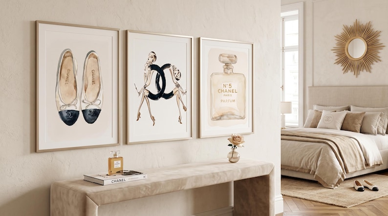 May include: Three framed watercolor prints of fashion items. The first shows a pair of ballet flats, the second a fashion illustration, and the third a perfume bottle. A small perfume bottle and a book are on a beige console table.