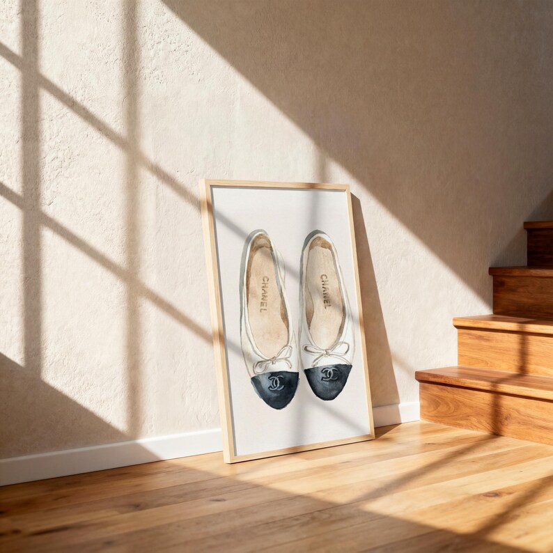 May include: Watercolor print of two ballet flats in a wooden frame. The shoes are white with navy blue toes and a bow detail. The word "Chanel" is printed on the side of each shoe. The artwork is displayed against a beige wall with sunlight.
