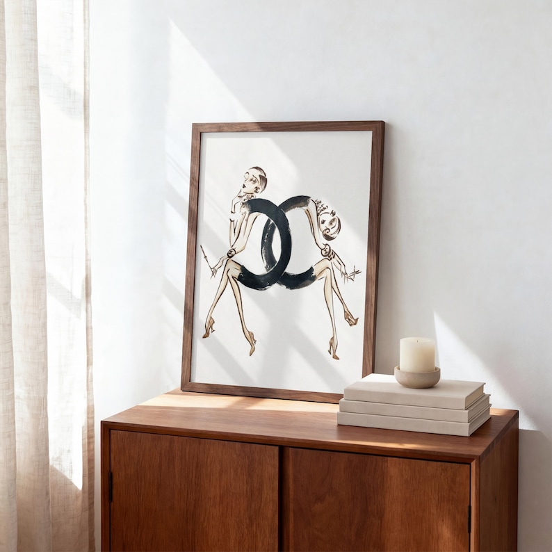 May include: A framed art print featuring a fashion illustration. The artwork depicts two stylized figures forming a black interlocking design. The figures are in a seated pose, with slender legs and arms. The frame is a dark wood color.