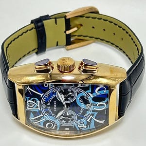 Puede incluir: Un reloj de pulsera dorado con forma de tonel, correa de cuero negro y hebilla dorada. La esfera muestra un patr&oacute;n de camuflaje azul y negro con grandes n&uacute;meros ar&aacute;bigos blancos, dos subesferas y el texto Franck Muller Geneve.