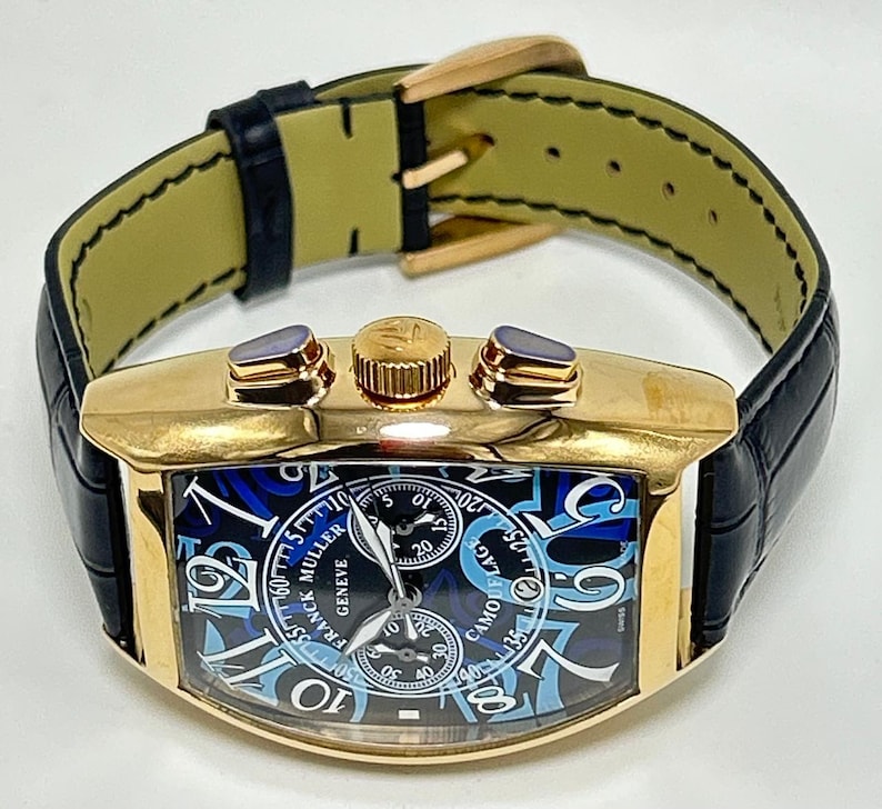 Puede incluir: Un reloj de pulsera dorado con forma de tonel, correa de cuero negro y hebilla dorada. La esfera muestra un patr&oacute;n de camuflaje azul y negro con grandes n&uacute;meros ar&aacute;bigos blancos, dos subesferas y el texto Franck Muller Geneve.