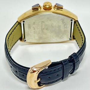 Puede incluir: Un reloj de pulsera dorado con forma de tonel, correa de cuero azul oscuro con relieve y hebilla dorada. El reloj presenta una tapa trasera met&aacute;lica con el texto grabado Franck Muller Geneve y B880 CC, junto con dos pulsadores dorados.