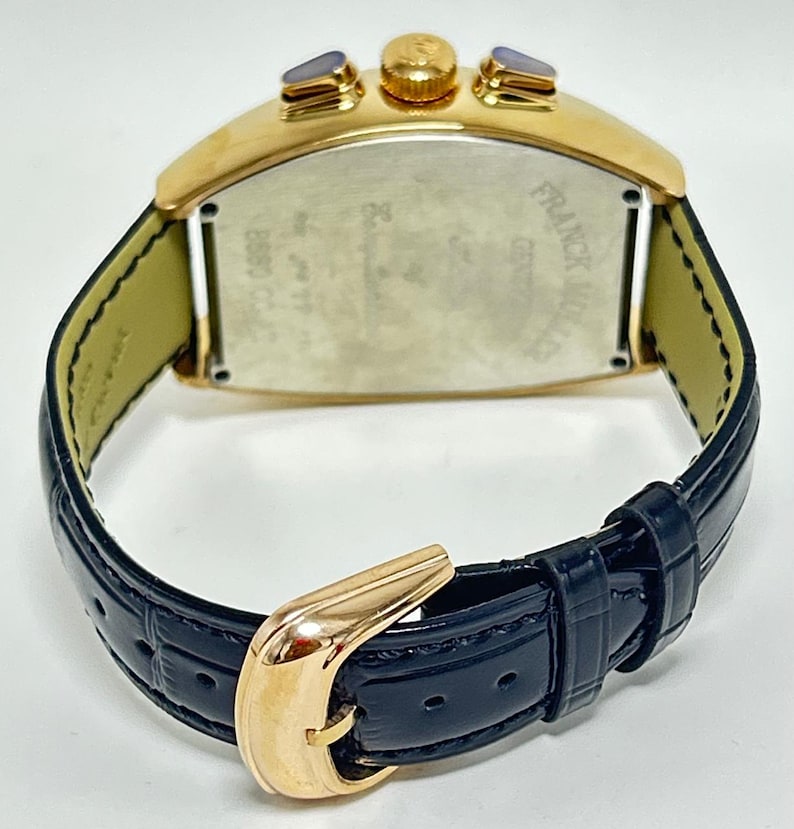 Puede incluir: Un reloj de pulsera dorado con forma de tonel, correa de cuero azul oscuro con relieve y hebilla dorada. El reloj presenta una tapa trasera met&aacute;lica con el texto grabado Franck Muller Geneve y B880 CC, junto con dos pulsadores dorados.
