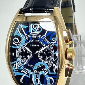 Puede incluir: Un reloj cron&oacute;grafo Franck Muller Geneve Camouflage con caja dorada en forma de tonel, correa de cuero negro y esfera negra con grandes n&uacute;meros ar&aacute;bigos azules, dos subesferas y una ventana de fecha en la posici&oacute;n de las seis.