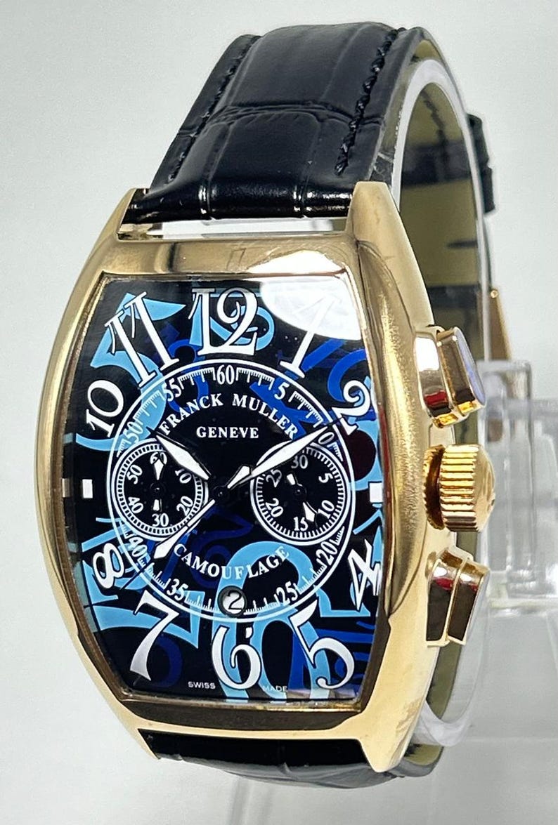 Puede incluir: Un reloj cron&oacute;grafo Franck Muller Geneve Camouflage con caja dorada en forma de tonel, correa de cuero negro y esfera negra con grandes n&uacute;meros ar&aacute;bigos azules, dos subesferas y una ventana de fecha en la posici&oacute;n de las seis.