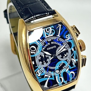 Puede incluir: Un reloj de pulsera dorado con forma de tonel y correa de cuero negro. La esfera presenta un patr&oacute;n de camuflaje azul, grandes n&uacute;meros ar&aacute;bigos blancos y el texto Franck Muller Geneve y Camouflage. Incluye tres subesferas y una ventana de fecha.