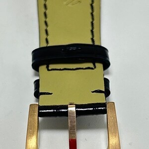 Puede incluir: Una correa de reloj con exterior negro brillante de acabado texturizado y reverso amarillo p&aacute;lido. La correa incluye una hebilla de metal dorado con un peque&ntilde;o logotipo grabado, dos trabillas de cuero negro y costuras negras en los bordes.