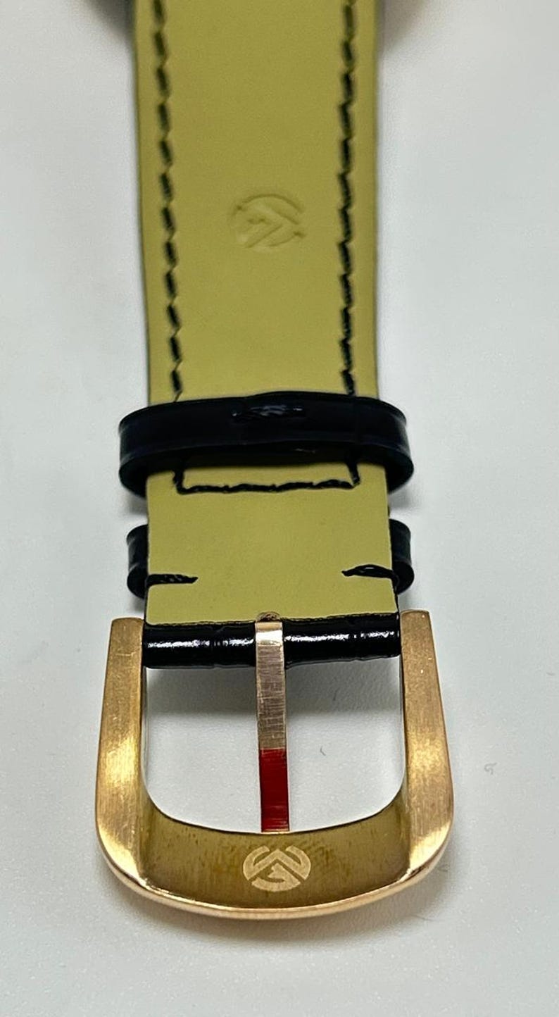 Puede incluir: Una correa de reloj con exterior negro brillante de acabado texturizado y reverso amarillo p&aacute;lido. La correa incluye una hebilla de metal dorado con un peque&ntilde;o logotipo grabado, dos trabillas de cuero negro y costuras negras en los bordes.