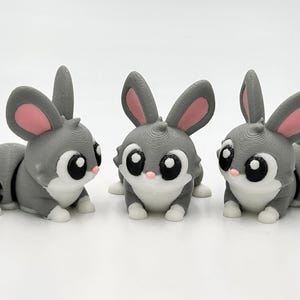 Peut inclure: Trois petites figurines de lapin articul&eacute;es dans des tons gris, blanc et rose. Chaque lapin pr&eacute;sente de grands yeux noirs avec des reflets blancs, l'int&eacute;rieur des oreilles rose, un nez rose et des pattes blanches, en position assise.