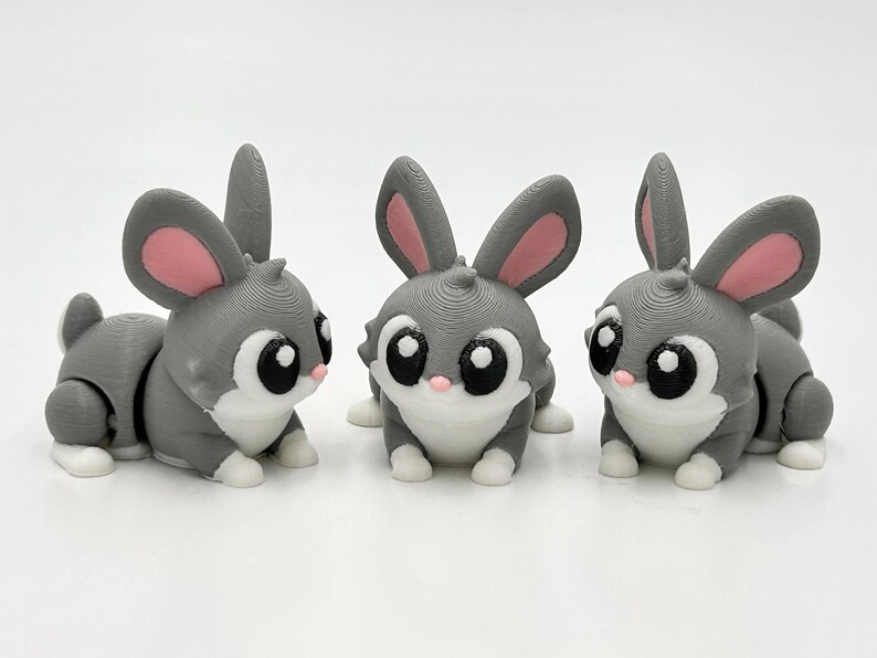 Peut inclure: Trois petites figurines de lapin articul&eacute;es dans des tons gris, blanc et rose. Chaque lapin pr&eacute;sente de grands yeux noirs avec des reflets blancs, l'int&eacute;rieur des oreilles rose, un nez rose et des pattes blanches, en position assise.