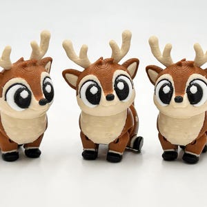 Peut inclure: Trois petites figurines de cerf marron et cr&egrave;me avec de grands yeux noirs et des bois clairs. Chaque cerf pr&eacute;sente une surface textur&eacute;e, des taches blanches sur le dos et de petits sabots noirs, align&eacute;s sur un fond blanc uni.