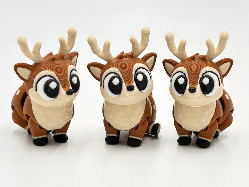 Peut inclure: Trois petites figurines de cerf marron et cr&egrave;me avec de grands yeux noirs et des bois clairs. Chaque cerf pr&eacute;sente une surface textur&eacute;e, des taches blanches sur le dos et de petits sabots noirs, align&eacute;s sur un fond blanc uni.