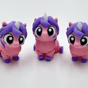Peut inclure: Trois petites figurines de licorne imprim&eacute;es en 3D avec des corps roses, des crini&egrave;res violettes et des cornes blanches. Chaque licorne a de grands yeux noirs et blancs, de petits sabots violets et une posture assise.